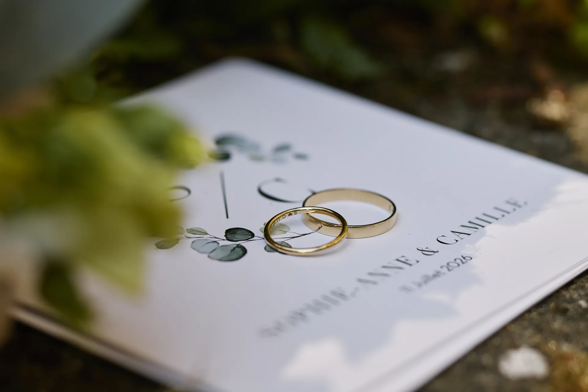 Deux alliances dorées posées sur un carton d'invitation de mariage avec des motifs de feuilles vertes