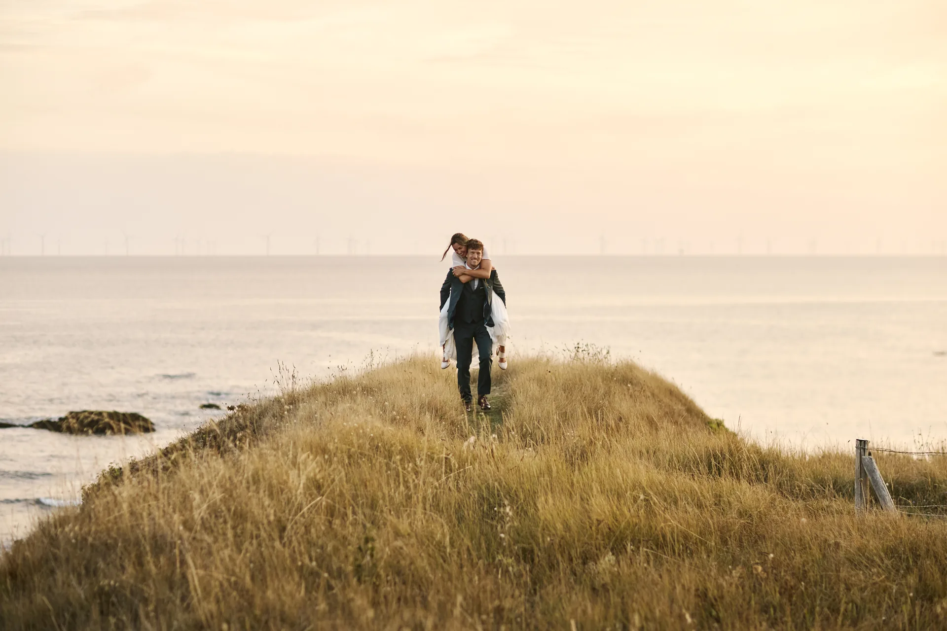 Un homme portant une femme en robe blanche sur son dos sur un chemin herbeux au bord de la mer au coucher du soleil