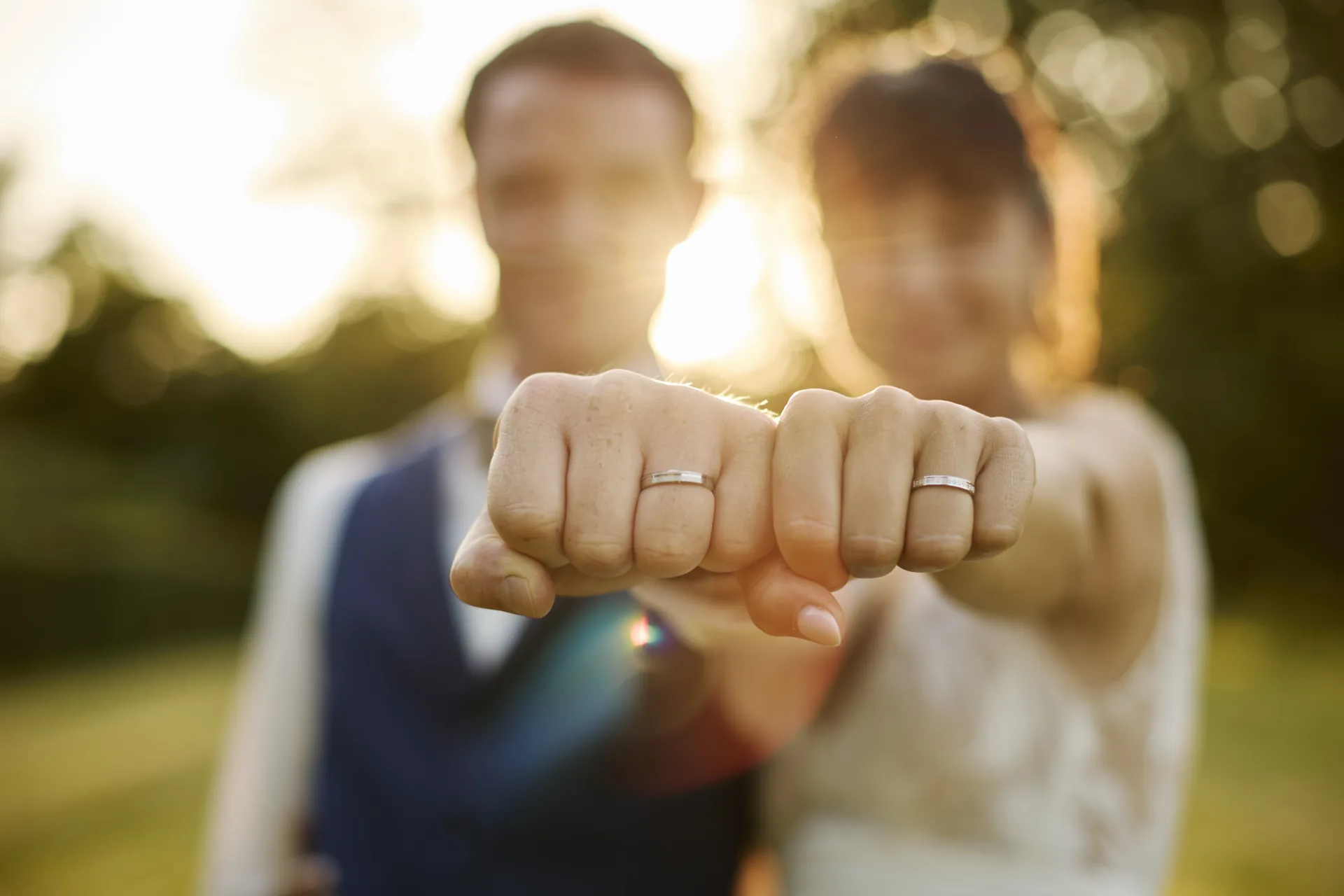 Un couple de mariés montre leurs alliances en serrant les poings vers la caméra avec un arrière-plan flou et un éclairage naturel
