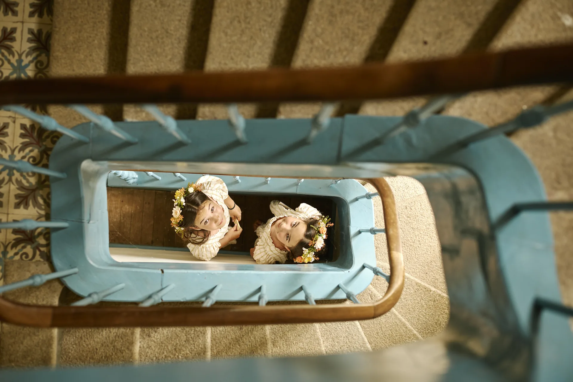 Deux petites filles en robes blanches avec des couronnes de fleurs regardant vers le haut dans une cage d'escalier bleue