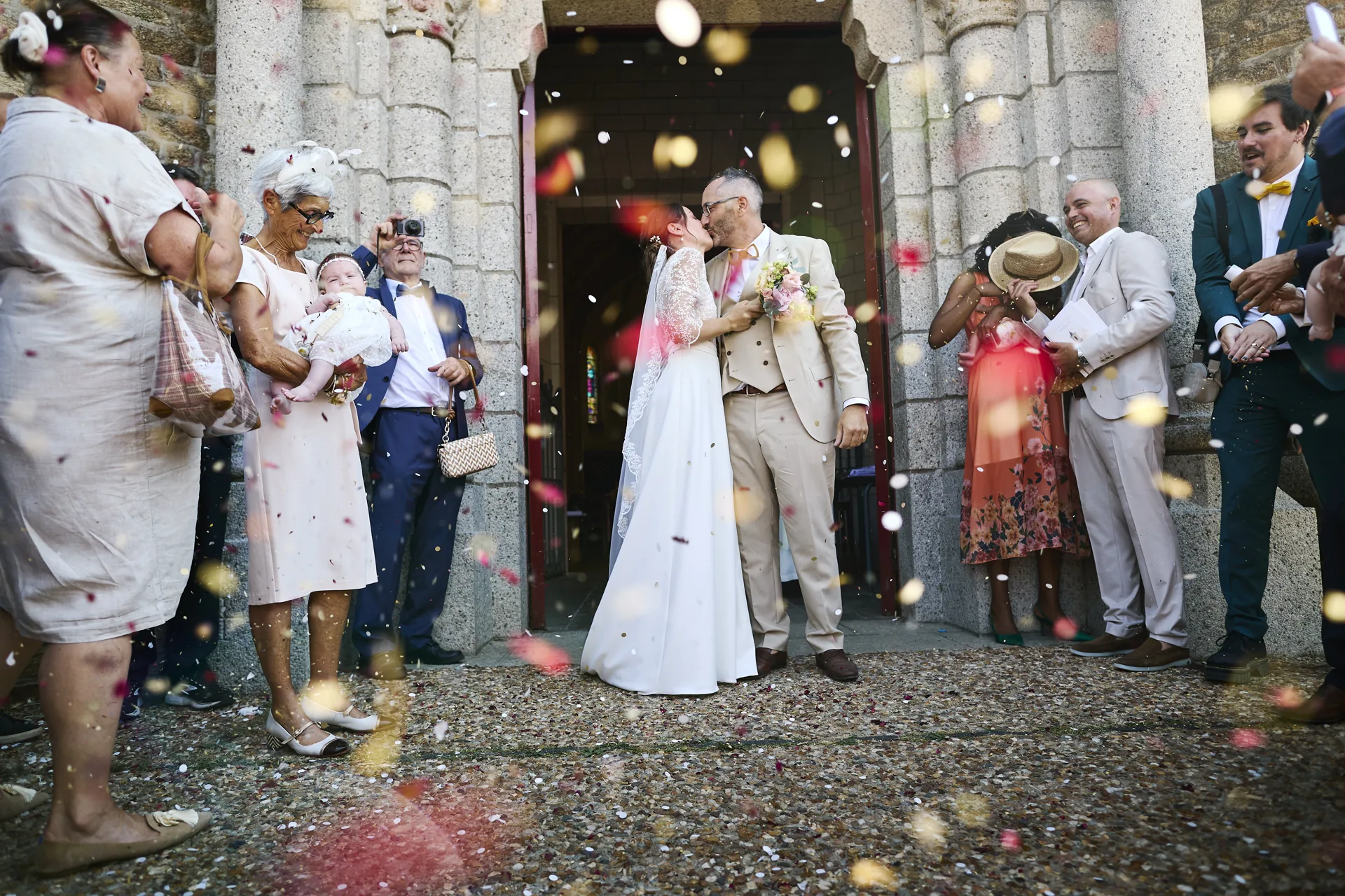 Un couple de mariés s'embrasse devant une église tandis que des invités jettent des pétales de fleurs colorés.