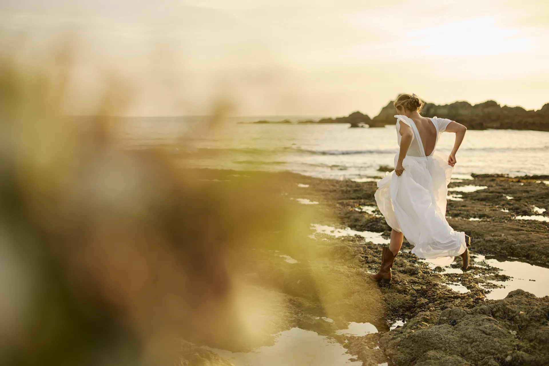Une mariée en robe blanche marche sur des rochers au bord de la mer au coucher du soleil