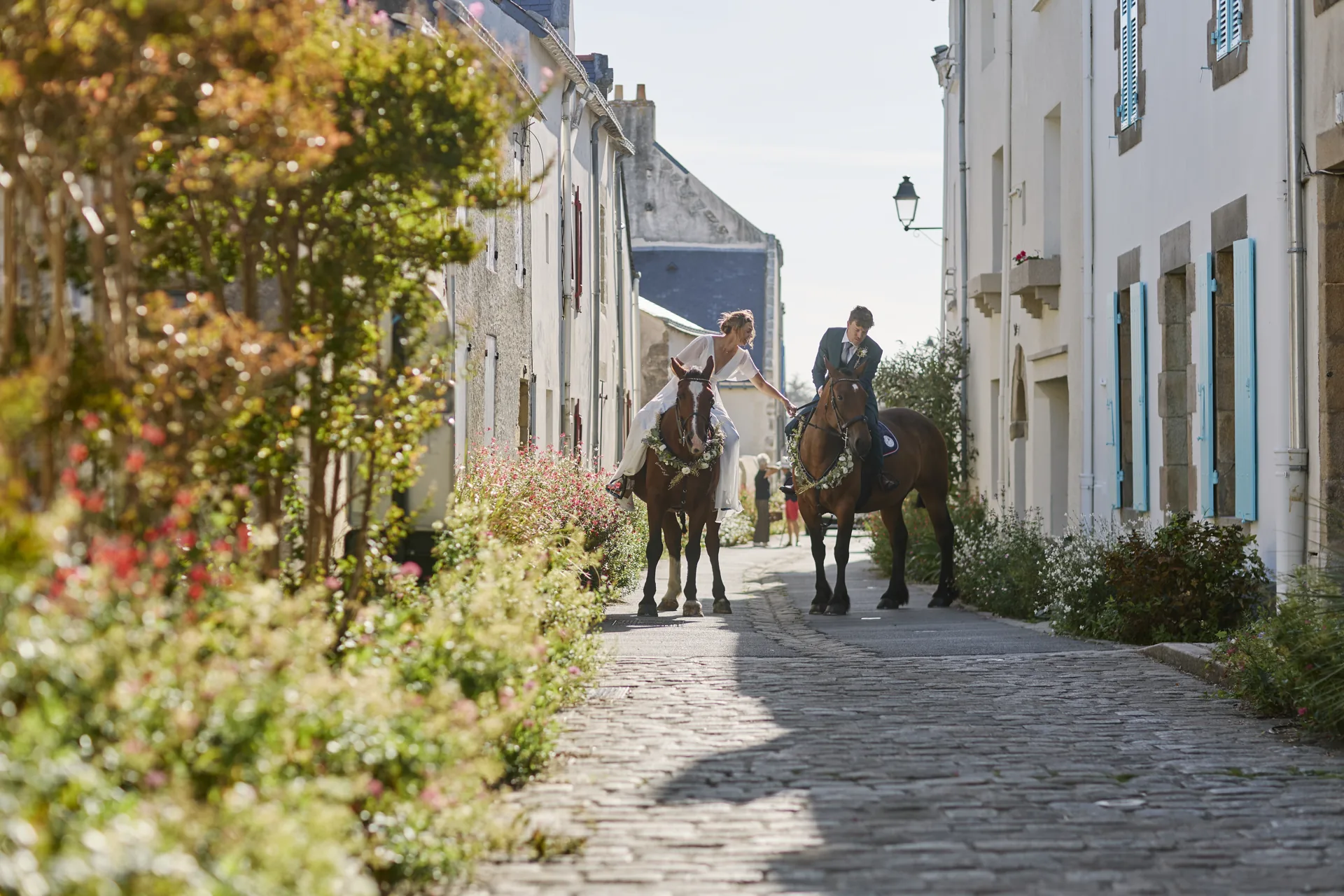 Un couple de mariés à cheval dans une ruelle pavée bordée de maisons blanches et de fleurs.